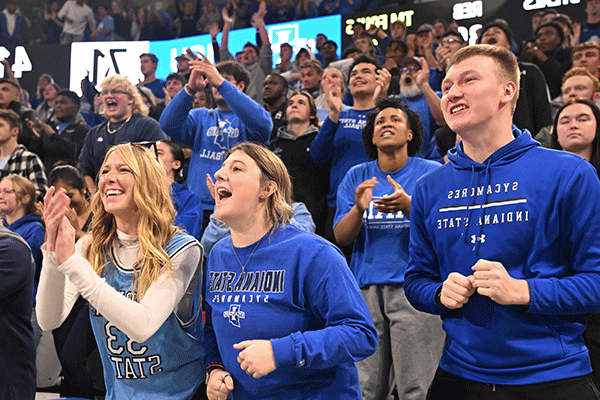 A diverse group of smiling Sycamore fans, most wearing various shades of blue Indiana State clothing, cheer and clap at a home Indiana State basketball game in the Hulman Center. Digital signage and a scoreboard are just visible behind them.